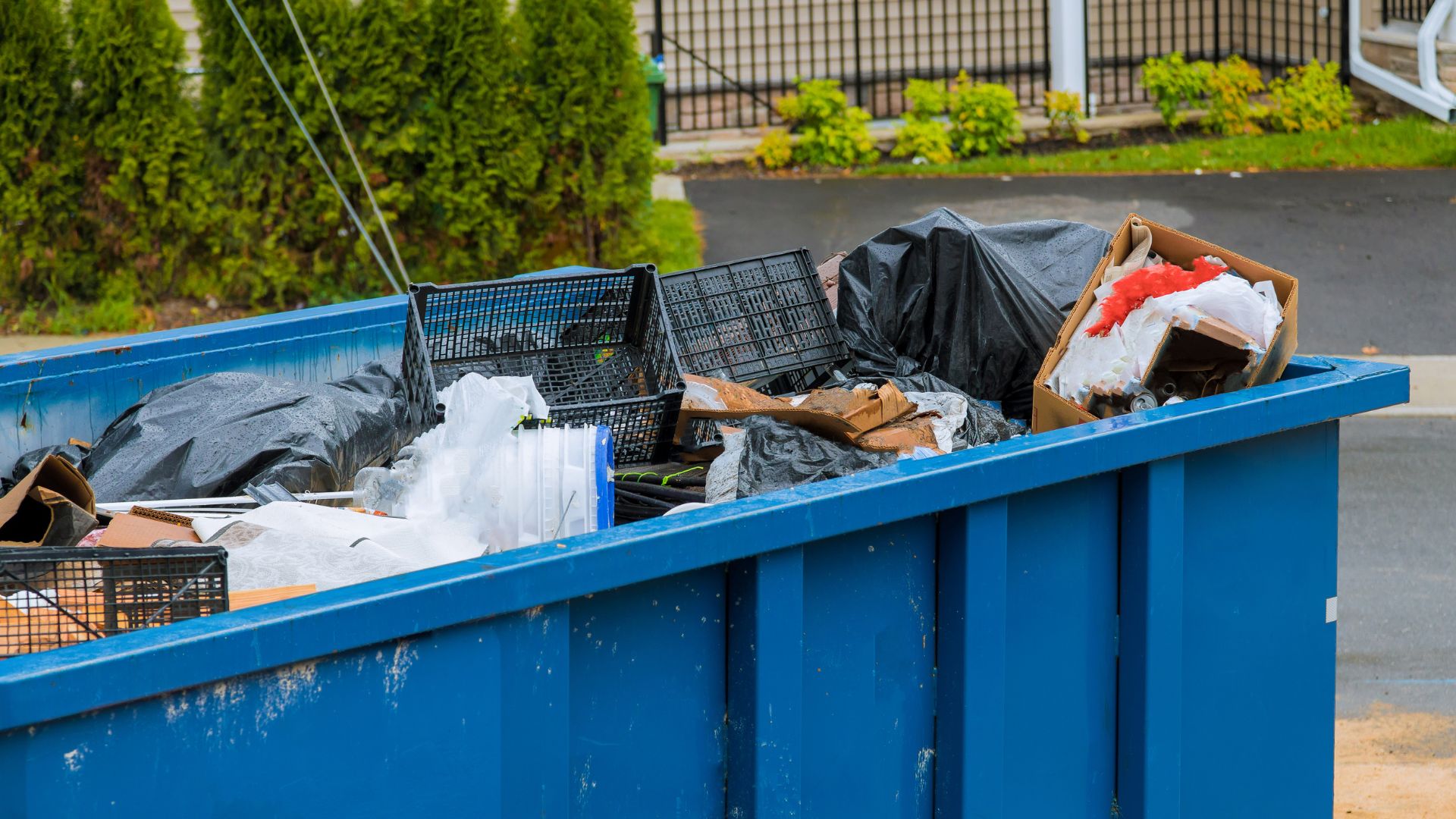 Blue dumpster filled with trash, plastic crates, and black garbage bags