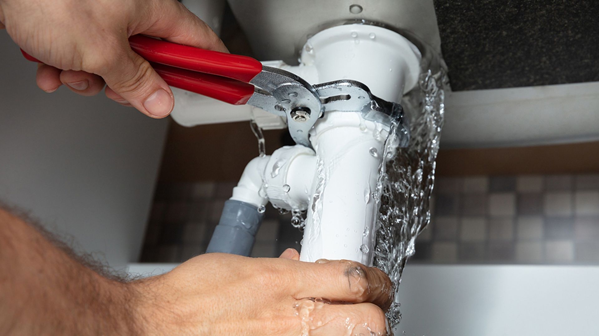 Plumber fixing a leaking white pipe with pliers and water spraying